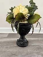 Front view of artificial flower bouquet with pale yellow flowers in a black carved wooden vase on a wood floor.
