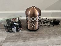 Bronze Better Homes & Gardens ultrasonic aroma diffuser with Moroccan scroll pattern, essential oils in zip bag, and power adapter visible behind on a wooden floor.