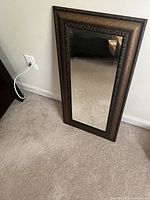 Mirror leaning against a white wall on carpeted floor showing dark bronze and gold shaded frame with beveled mirror surface.