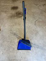 Front view of the lobby broom and blue dustpan set placed on a concrete floor, showing broom with missing top cover.