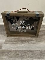 Front view of wooden fund bank showing glass front with 'Our Adventure Fund' text and world map behind glass, leather-like handle on top, metal corner accents.