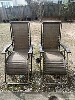 Front-facing view of two tan zero gravity lounge patio chairs on outdoor grass showing metal frames and fabric seats.