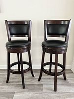 Front view of two swivel bar stools with black leather seats and backs, dark wood frames, placed side by side on wooden flooring.