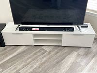 Front view of white TV stand with a large flat screen TV on top, showing closed drawers and open shelves with a soundbar.