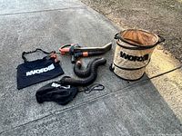 Full view of all items included in the lot laid out on concrete. Items include the WORX electric leaf blower, collapsible yard waste bag, debris collection bag, flexible hose, and carrying strap.