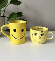 Yellow tall smiley face mug and short mug with 3D nose and 'Good Morning' text, placed side by side on a white surface with greenery in background.