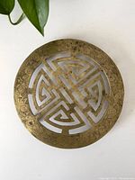 Top-down image showing circular brass trivet with intricate geometric cut-out design and ornate border etching.