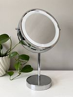 Front view of the round double-sided chrome metal vanity mirror with built-in light ring, standing on round base, with green plant in background.