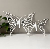 Two white wicker butterflies against a grey background, showing overall design and size difference.