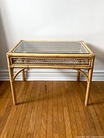 Full view of the square vintage wicker bamboo table with a clear glass top. Shows the overall structure and bamboo frame against a white wall and wooden floor.