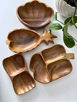 Photo of four uniquely shaped vintage wooden bowls including scalloped round, pineapple, divided section, and scalloped oval bowl, arranged on a white surface with green leaves in background.