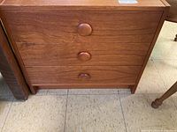 Front view of wooden bedside table showing three drawers with round wooden knobs and warm brown wood grain finish.