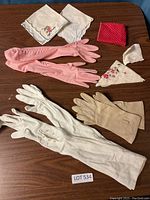 Top view of multiple gloves and embroidered handkerchiefs laid on a wooden table including white silky gloves, beige suede gloves, long pink gloves, and several handkerchiefs with floral embroidery and one red polka-dot.