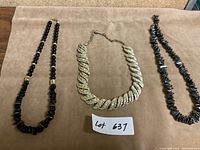 Three necklaces displayed on a beige surface: a black beaded necklace with gold accents, a thick silver-tone twisted rope chain, and a hematite chip bead necklace.
