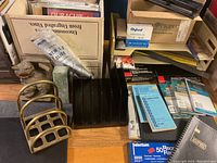 Photo showing a black metal desktop organizer with vertical slots, brass letter/file holders, packages of binder clips, file labels, and office supplies on a desk.