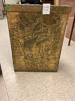 Front view of gold-toned, textured metal umbrella stand with embossed Asian-inspired boat and tree design.