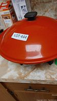 Orange electric wok lid with black knob on counter, showing lot number sticker.