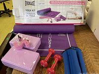 Everlast yoga kit items displayed including purple yoga mat rolled out, two purple yoga blocks, yoga strap, and pink dumbbells with their original box in background.