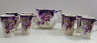 Full view of set featuring one jug and eight matching mugs decorated with grape pattern on white porcelain.