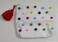 Front view of a white 100% cotton bag with multicolor pom poms and red tassle attached to zipper