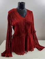 Front view of red pleated blouse displayed on mannequin, showing long sleeves, V-neck, and flared cuffs.