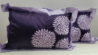 Two purple/lilac rectangular decorative cushions displayed front and slightly angled to show floral pattern and border details, zipper closure not visible.