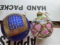 Close-up of two decorated glass Christmas ornaments, one blue with gold and glitter details, and one clear with pink hearts and gold lattice.