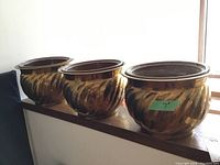 Three 7 inch brass pots with swirled exterior design lined up on a shelf with dark ceramic liners inside