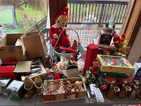 Overview of assorted Christmas decorations on table including elf figures, boxes of ornaments, candle holders, tinsel and small figurines