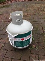 White propane tank with green danger label and metal top handle, placed on brick surface.