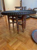 Carved wood folding table base supporting round brass tray on parquet floor.