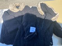 Photo showing two black textured coats laid side by side, both with light beige fur trim collars, one fully open and one folded.