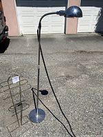 Mid-century metal floor lamp and brass rack photographed outside on pavement in front of garage doors, showing full height and structure of lamp and rack.