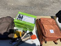 Overview of lot showing unopened disposable dinnerware set, brown wheeled suitcase, brown backpack and camping utensils on white table outside on pavement.