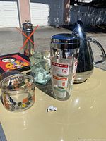 Photo showing the cocktail shaker with cocktail recipes, the metal pitcher, clear glass bowls, and clear glass bowl with floral design alongside other small items on a glass table outdoors.