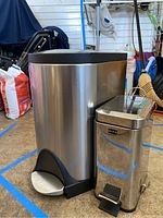 Two silver metal trash cans of different sizes side by side in a garage with visible foot pedals and black lids.