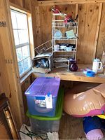 Photo of a wire storage rack on a wooden table with miscellaneous items including desk lamps, painting supplies, and plastic bins below the table.