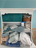 Full view of white shelving unit holding fluffy towels in various colors and textures, teal shag bath mat, and wicker basket with rolled towels.