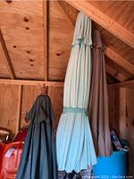 Three large outdoor umbrellas in dark green, light blue, and brown, folded and hanging inside a wooden shed.