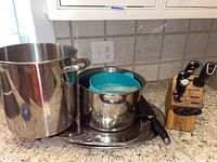 Overview of large and smaller stainless steel pots with blue-green silicone utensil rests inside smaller pot, served on stainless steel platters, next to Wusthof wooden knife block with knives