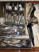 Drawer with multiple compartments containing Reed and Barton stainless steel flatware including forks, spoons, and knives along with kitchen utensils.