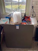 Brown square card table with household supplies placed on and around it, items visible include bubble wrap rolls, Pampers wipes box, trash bags, dust pans, paper towel dispenser.