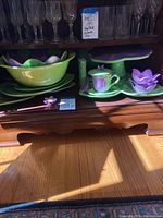 Suzanne Somers Violets hand painted serving bowls and matching platters in green and purple floral motif displayed inside a wooden cabinet shelf.