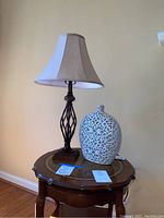 Full view of the wood end table with glass inset, ceramic vessel, and table lamp on top, against a beige wall and wooden floor.