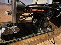 Close-up front and right side view of black Commander sewing machine on oak cabinet base with visible spool of white thread.