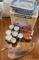 Overall view showing Breadman Ultra Breadmaker box, wooden breadboard shaped like a bird, ten assorted coffee mugs in front, and two aluminum foil baking pans stacked on top.