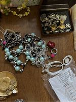 Close view of knotted necklaces with blue faux pearls, black and silver chain links, pink and red accent beads, and a small box holding large clear faceted beads and a gold-tone floral brooch.