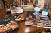 Photo showing multiple boxed Pampered Chef microwave cookware items including muffin pan, baking stone, apple corer/peeler, along with vintage cookbooks and other kitchen accessories.