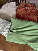 Four folded bedspreads and pillows laid out on a wooden table. Includes green striped pillow, orange velvety pillow, patterned gray pillow, and a light beige bedspread.