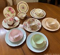 Photo showing full set of eight vintage teacup and saucer pairs on wooden table with floral and pastel designs.